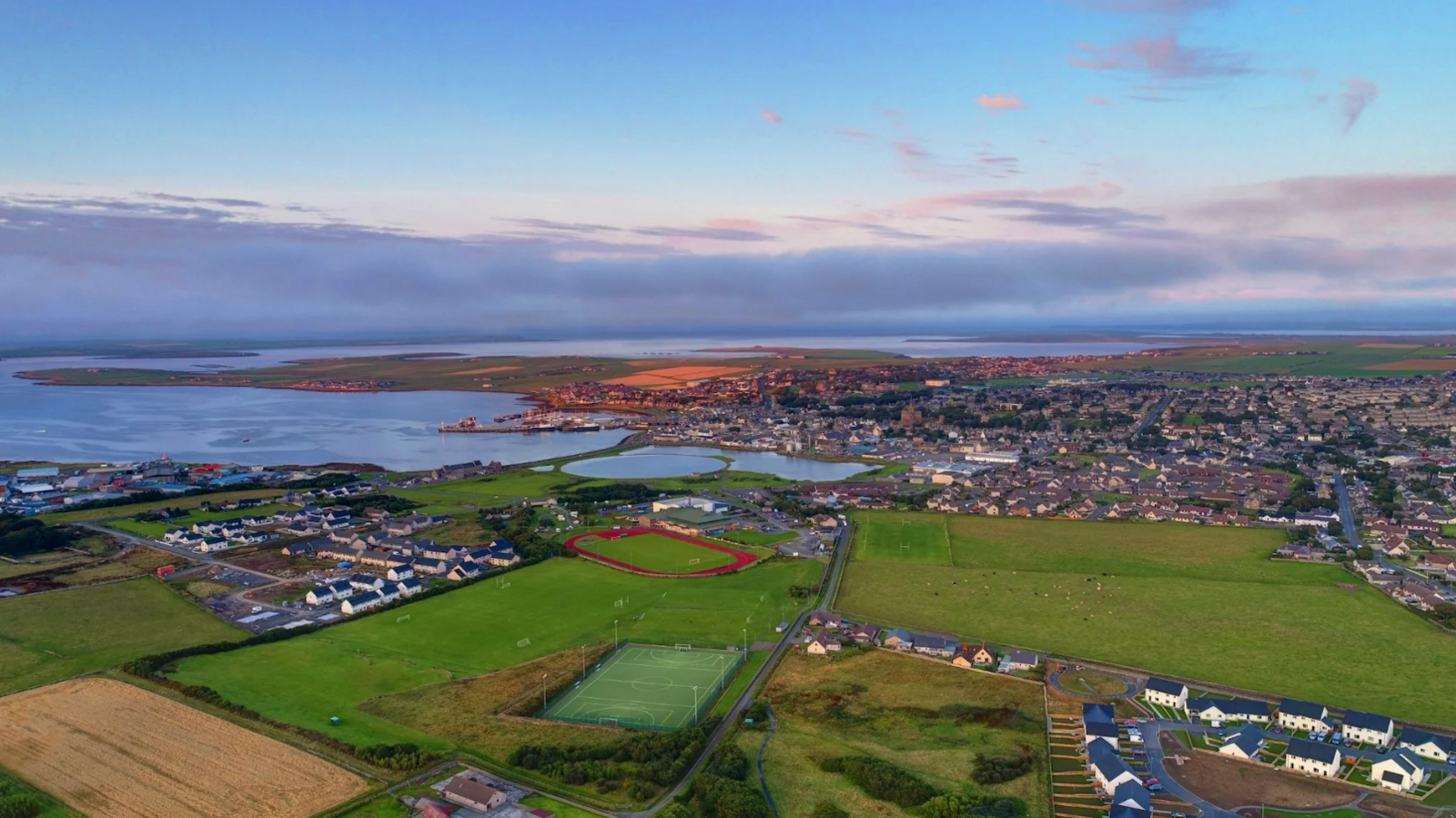 An aerial view of the Picky in Orkney. The sky is blue and grey. A number of green fields and houses are below. A running track surrounding a green sports pitch can be seen in the distance.