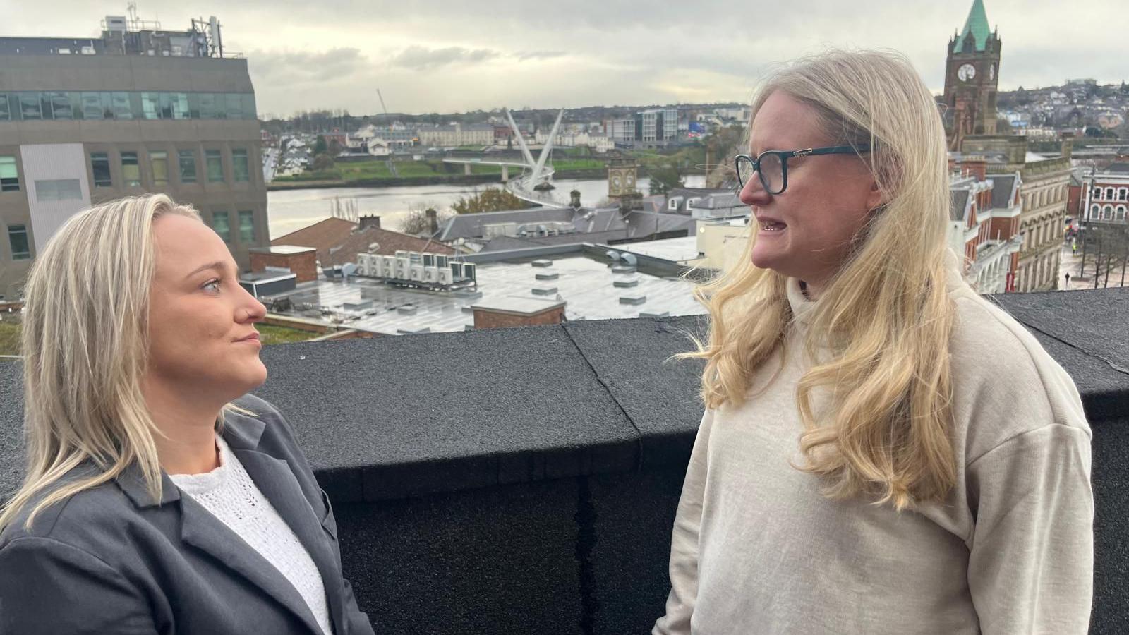 chloe wilson and gerladine hanna stand on the roof of a building overlooking derry's river foyle. Chloe is on the left . the city's peace bridge can be seen in the background, as can the guildhall and other buidlings. in the distance is river foyle and the waterside area of derry. chloe is wearing a grey jacket and whilte blouse, and has long blonde hair. geraldine on the right is wearing a white top, and also has blond hair. she is also wearing eye glasses.