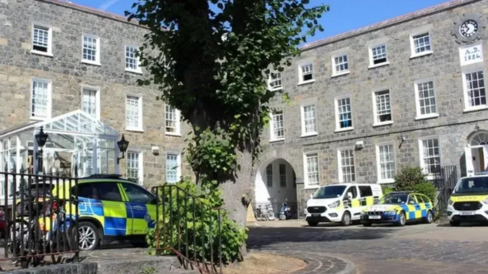 Guernsey Police headquarters - two buildings in an L shape with grey and brown bricks, white panelled windows and four police cars parked outside, a large tree in the middle, on a sunny day 