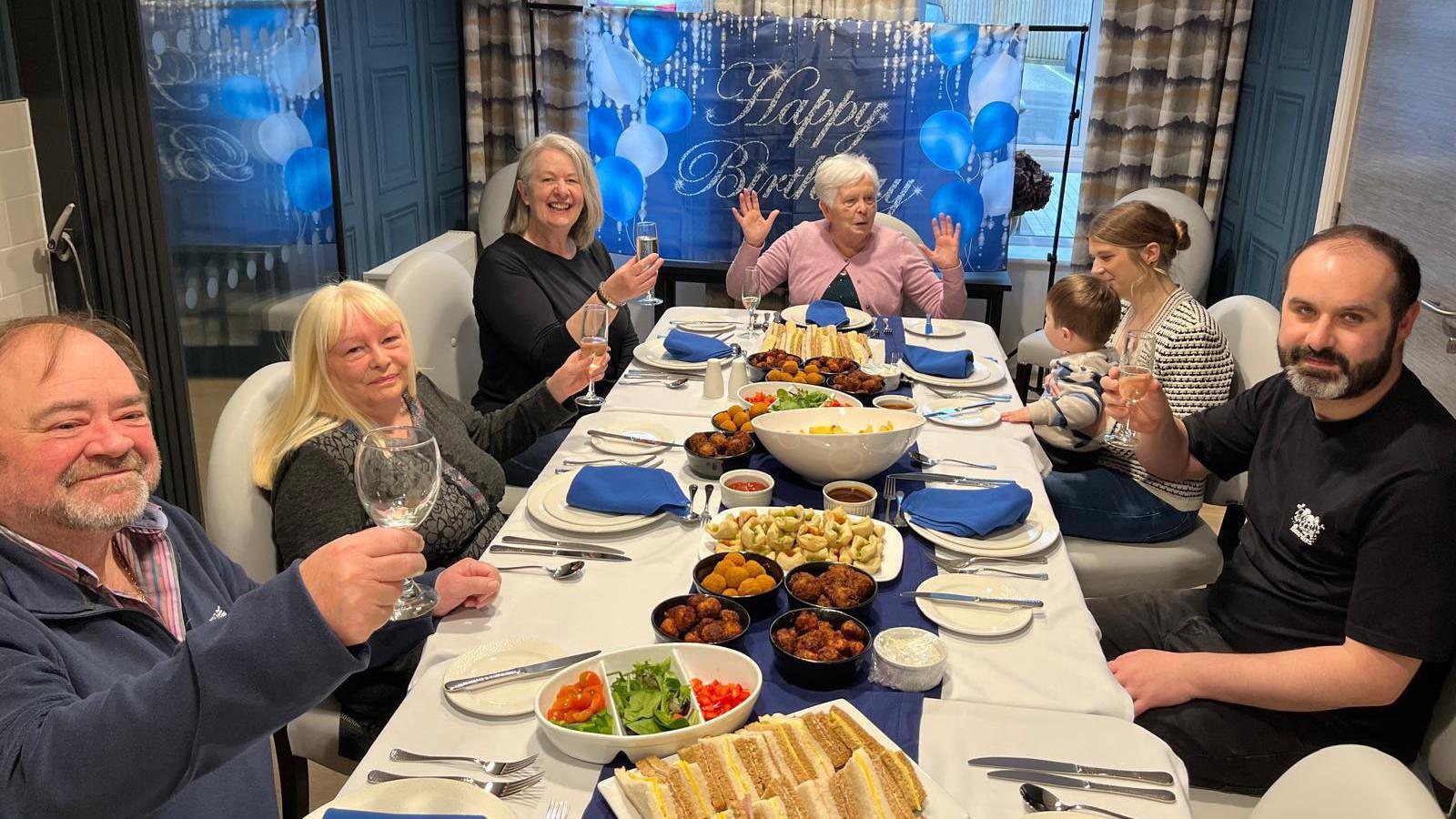 Barbara Wild sits at the head of the table as residents have a birthday lunch. Mrs Wild has her hands up as she talks to a woman with a child on her knee, and the other people are raising their glasses and looking at the camera.