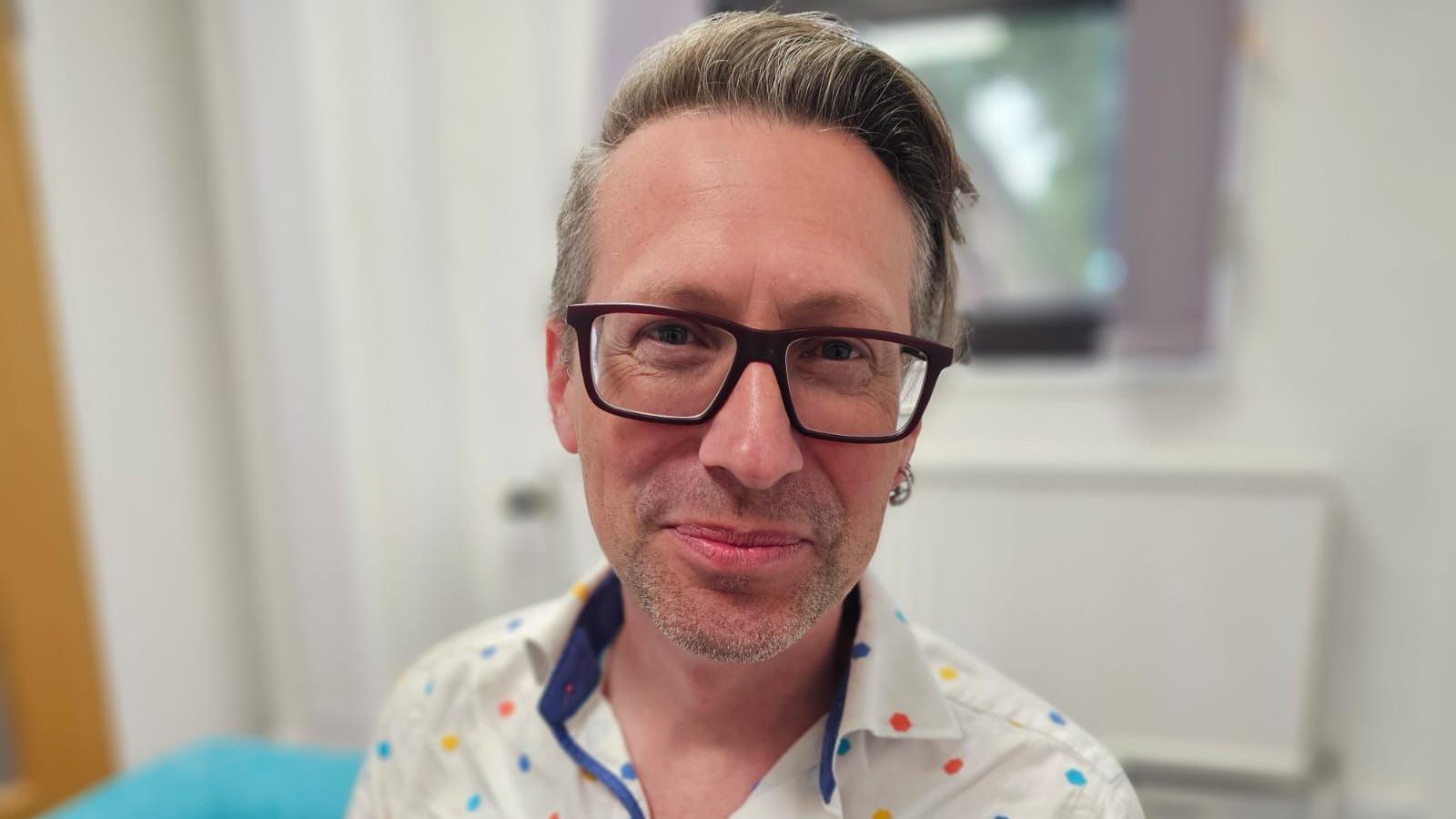Apollos is wearing a polka dot shirt and black-framed glasses. He has light grey/brown hair and is smiling at the camera.