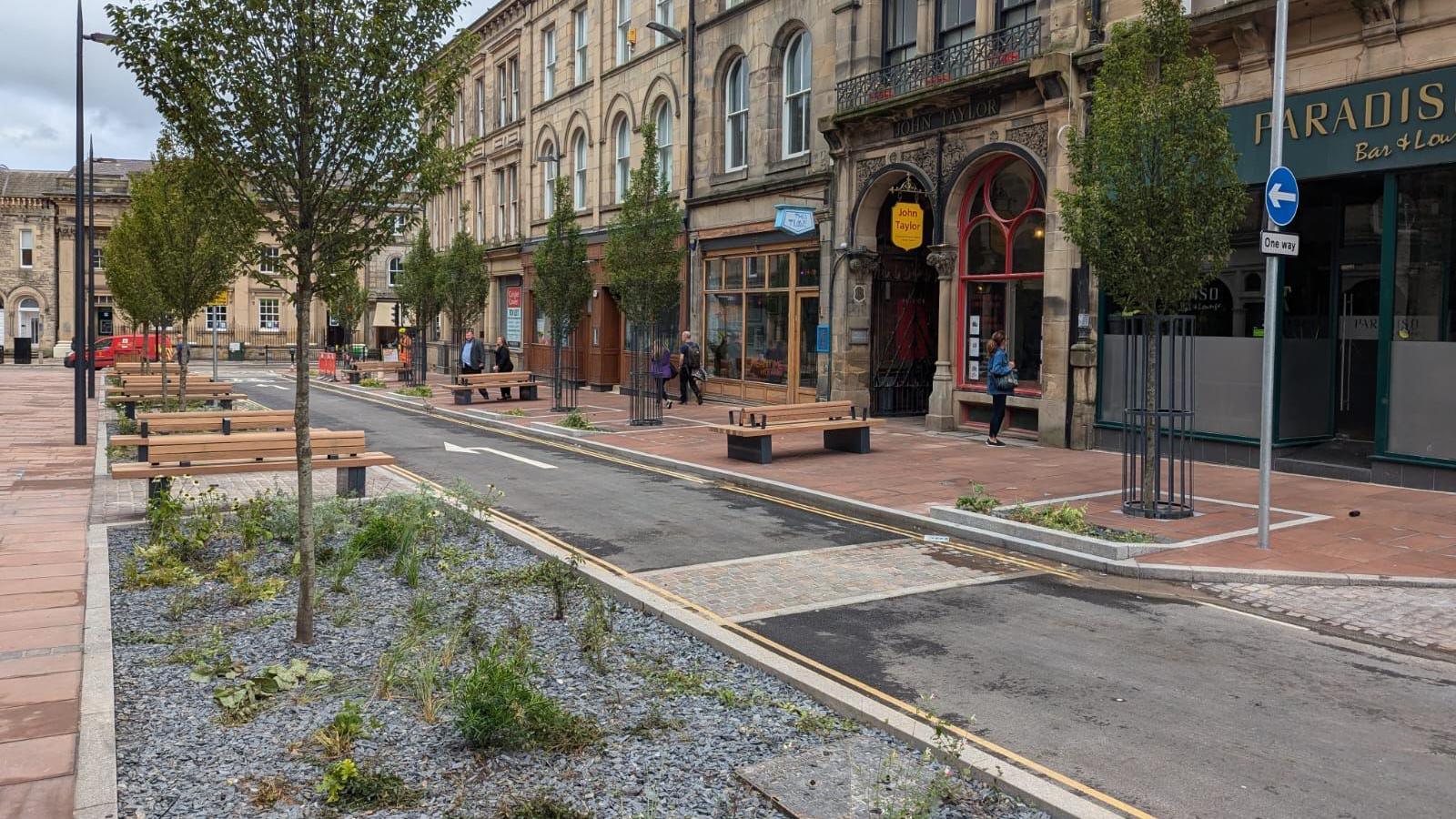 Devonshire Street after improvement work. It has wide pavements, with a single road, benches and flower beds in front of businesses. 