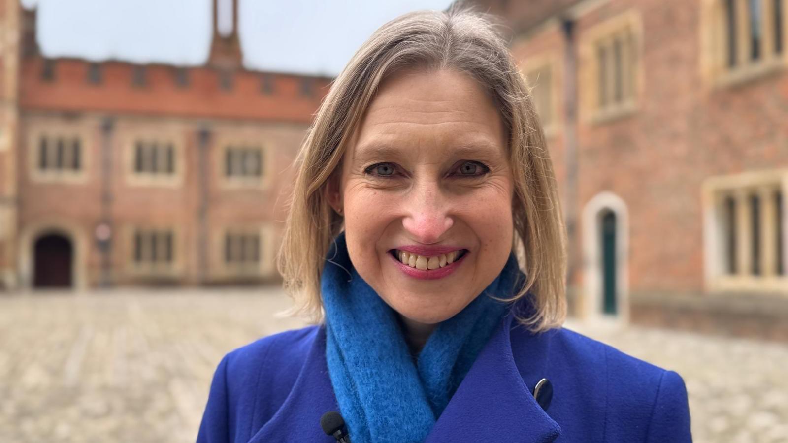 A head-and-shoulders photograph of Tracy Borman standing in a Tudor courtyard at Hampton Court Palace. She is wearing a blue coat with a light blue scarf and has shoulder-length blonde hair