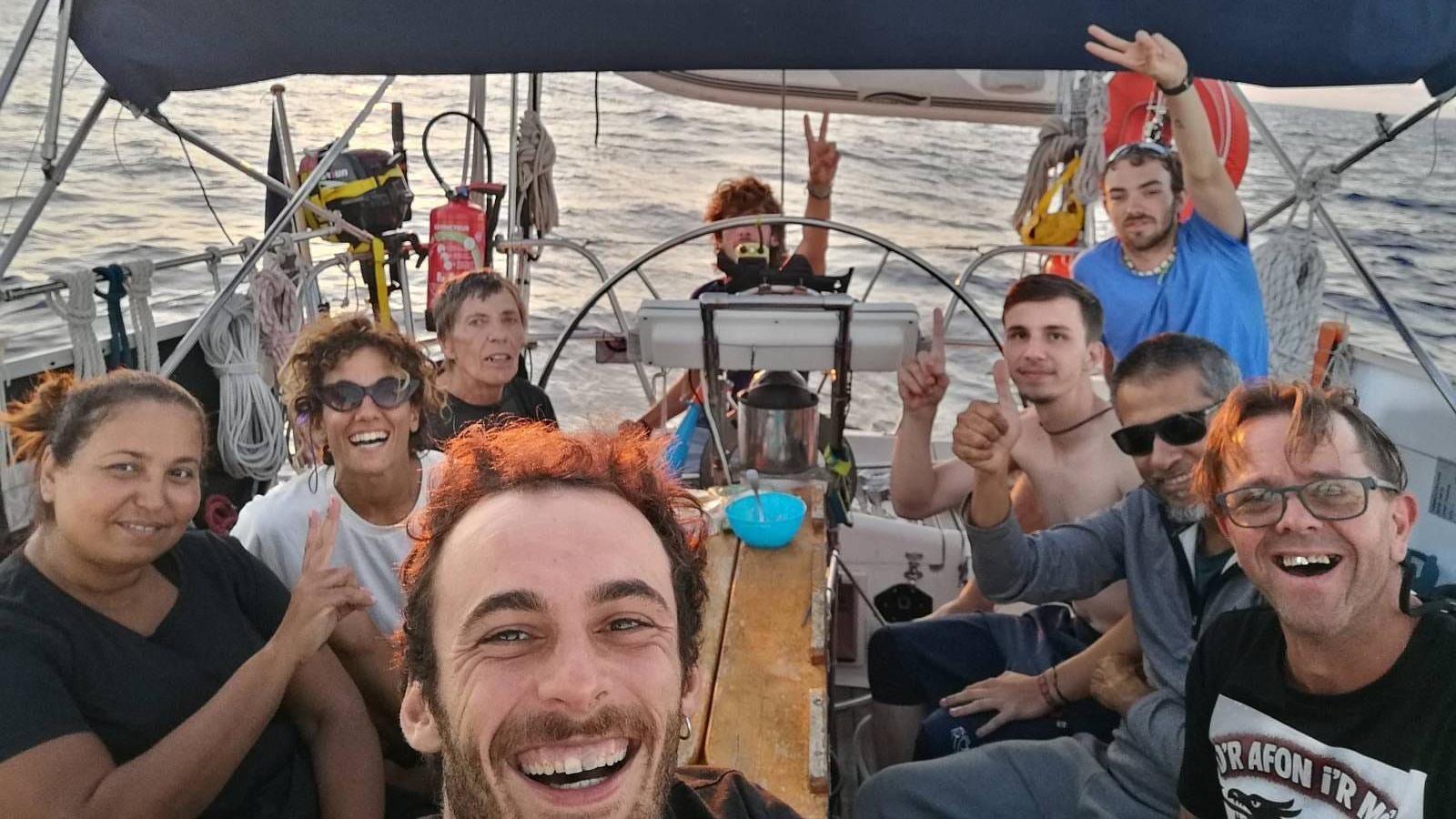 Eight people sit around a small table on a small boat. One of them is taking a selfie with a camera and they are all smiling. Some of them have their thumbs up while others are making peace signs. 