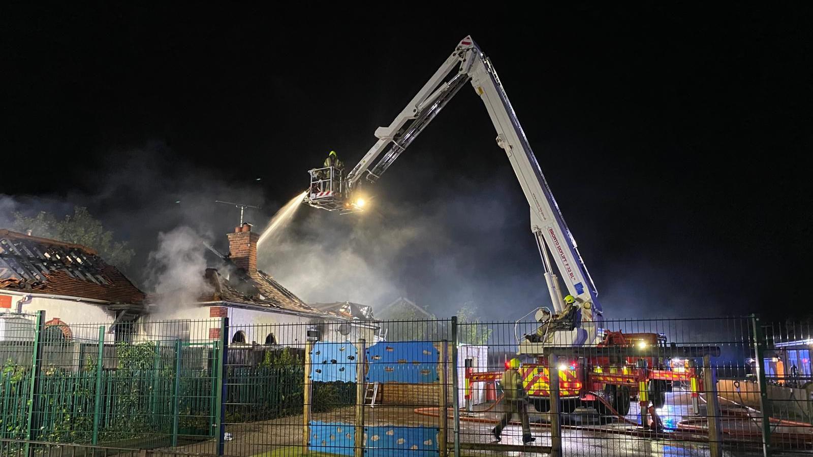 Firefighters on an aerial ladder are tackling a fire in a detached bungalow. Part of the roof is destroyed. Other homes can be seen on either side of the bungalow. There is a fenced off area at the front and other firefighters can be seen there
