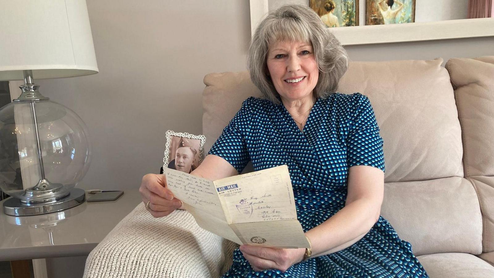 A woman with grey, white hair to her shoulders, sits in a light grey sofa with a table lamp to her right and a window behind her head. She wears a dark blue dress with light blue spots and holds a piece of folded paper in her hand with blue writing.