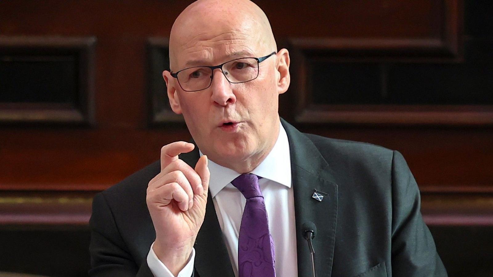 John Swinney making a speech - he is wearing a black blazer, a white shirt and a purple tie