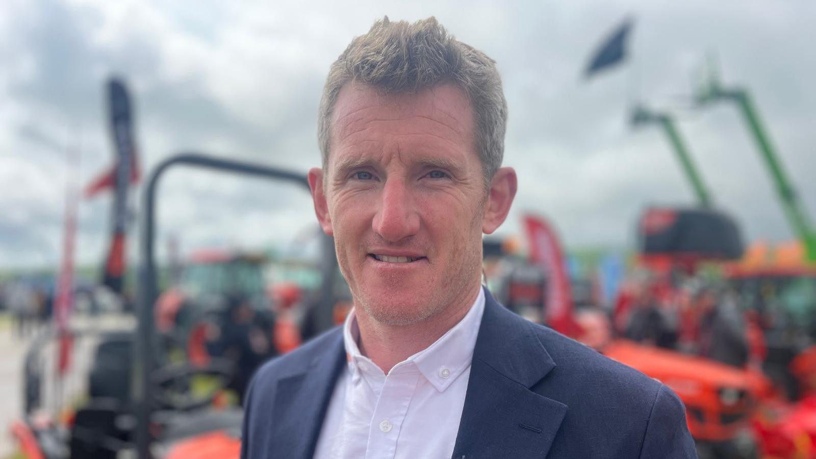 Man in suit jacket and open shirt standing in front of farm machinery