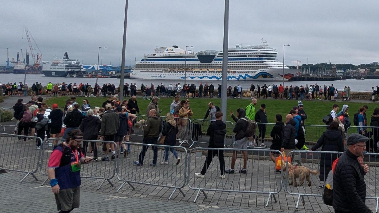 Crowds of people walking in metal barriers towards a ferry terminal. In the background is a larger holiday ferry, as well as the smaller Shields Ferry on the other side of the River Tyne.