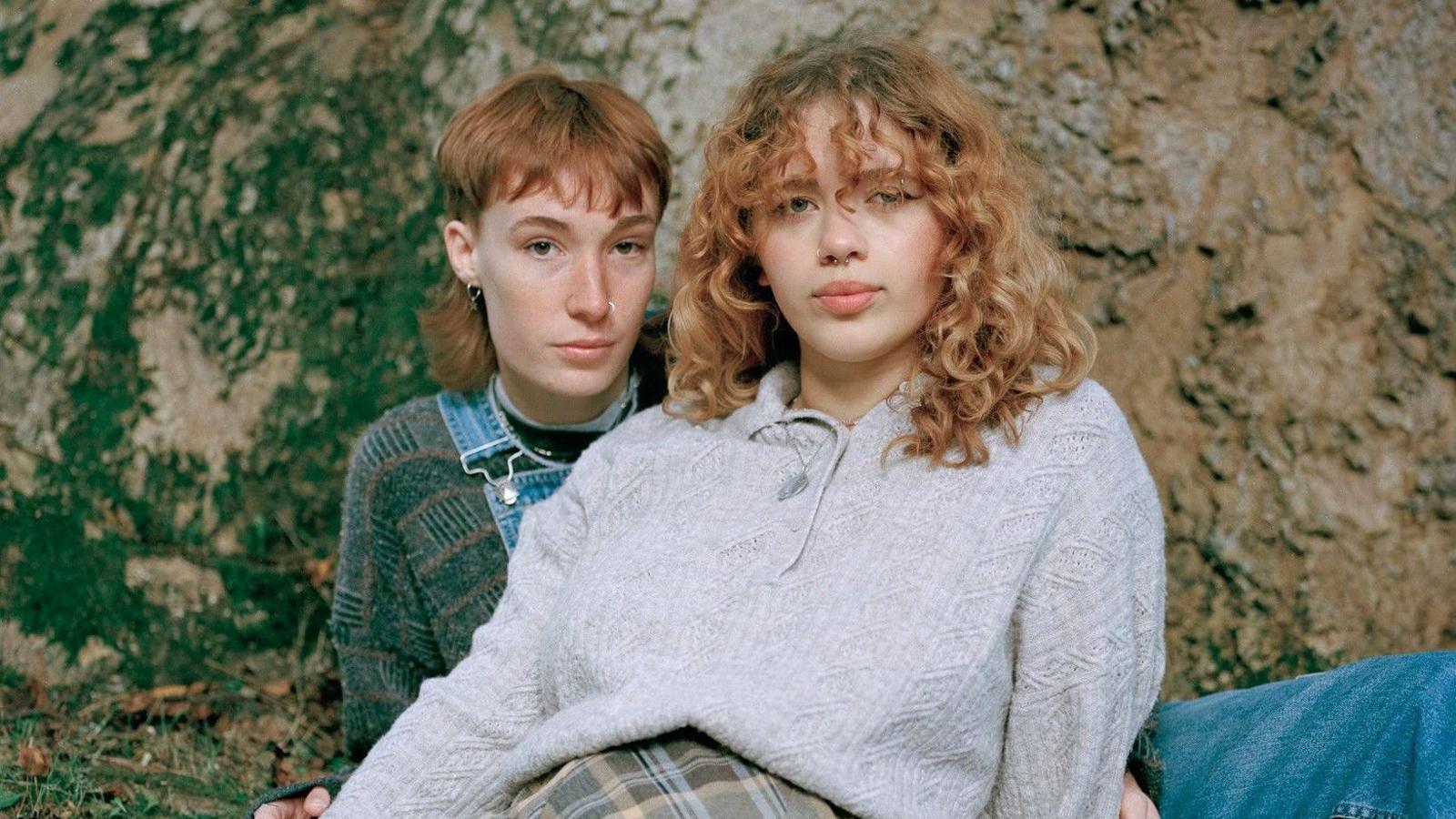 Image of the winning portrait called Maria and Connie. It shows two people laying closely together. The pair have light hair and are both wearing knitwear. They are in front of what appears to be a rockface.