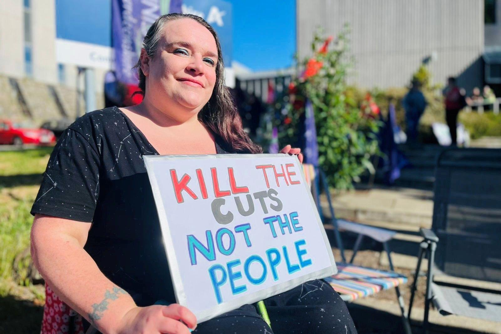 Woman holding a sign which says 'kill the cuts not the people'.
