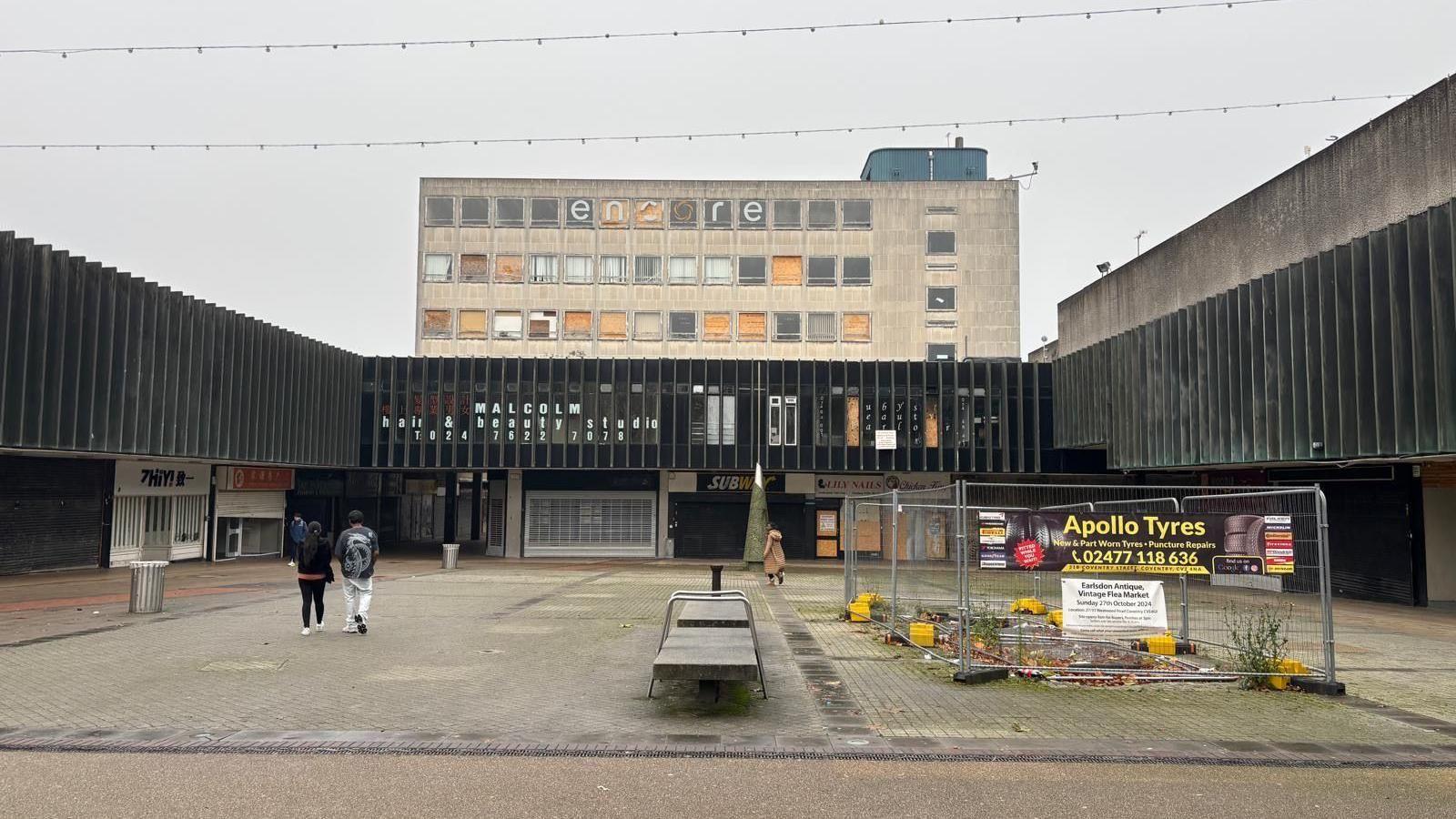 An old shopping precinct, almost deserted with just a few people walking across it. The few shops that are visible appear to be closed and the whole scene appears rather desolate.