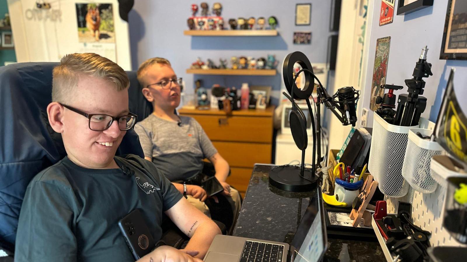 Sam and Alex in their bedroom. Alex is on a laptop and Sam is sat next to him. They are around a desk where filming equipment can be seen. 