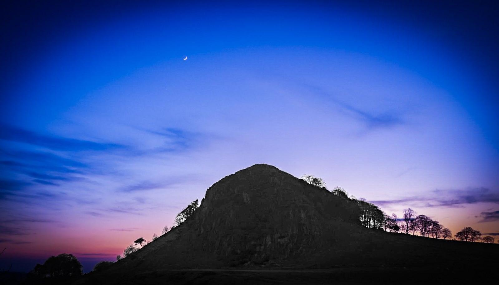Landscape image of the shadow of a hill and trees leading down with the vivid background of a blue, purple, and pink ombre sky and crescent moon