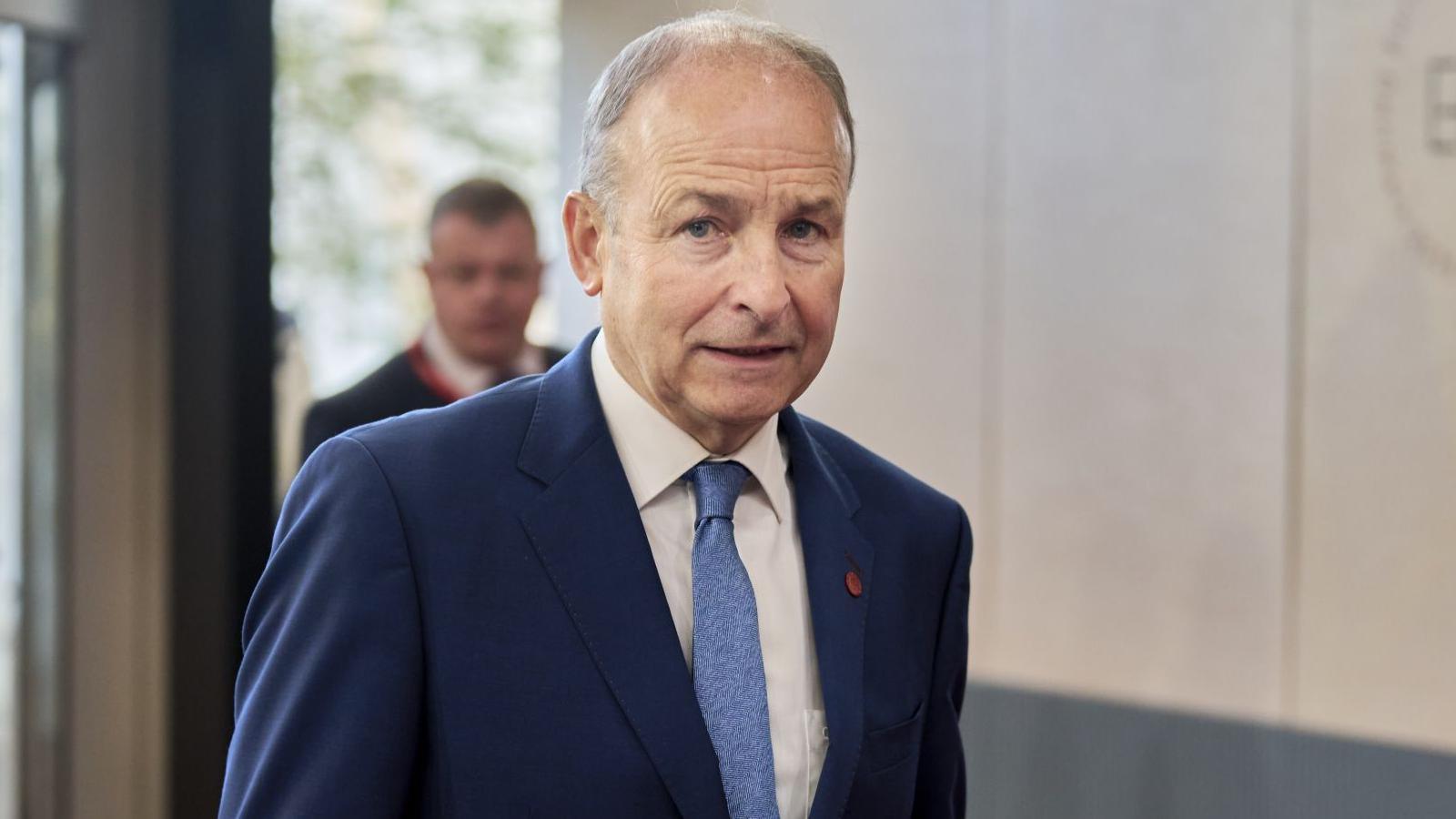 Micheál Martin looks at the cameras as he walks into a building in Copenhagen. He is an older man, with balding, short grey hair. He has a neutral expression on his face. He is wearing a navy suit jacket, a pale blue tie and a white collared shirt.