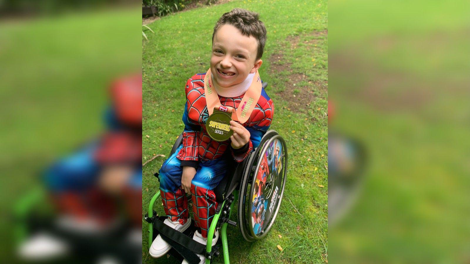 A young boy wearing a Spiderman costume while sat in a wheelchair. He is smiling at the camera and holding a gold-coloured medal which is around his neck