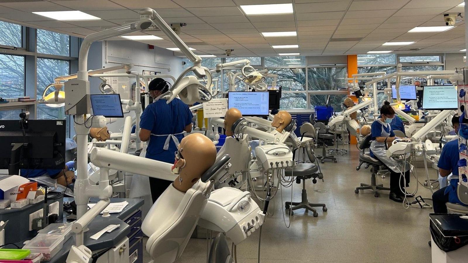 Dental students dressed in blue tunics practicing in a large room on phantom heads