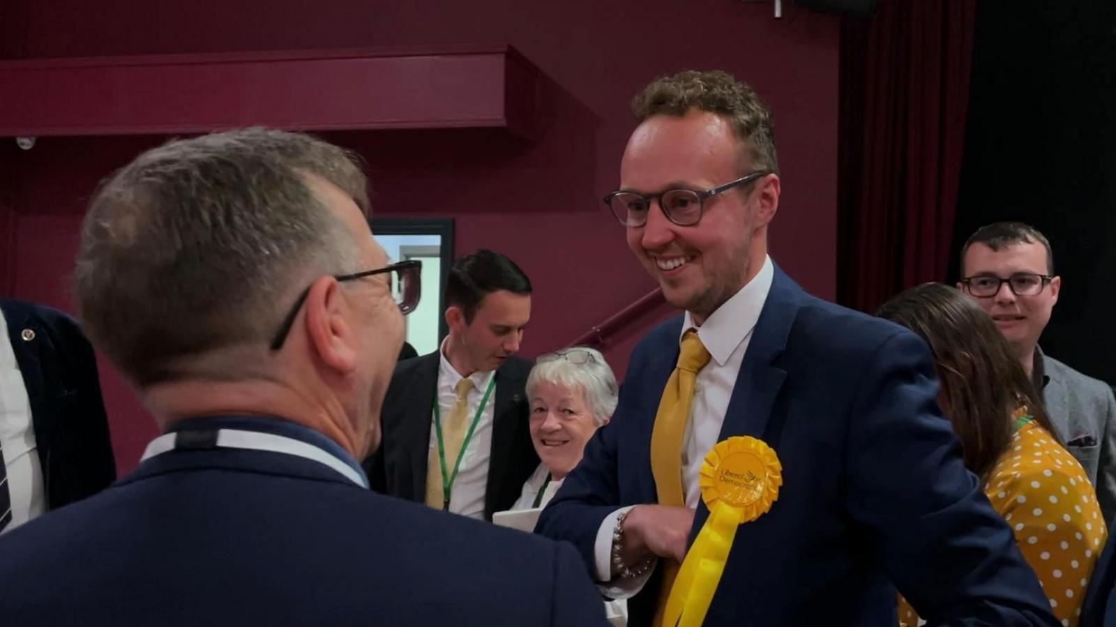 MP Adam Dance greets supporters while wearing a navy suit with a yellow rosette and tie