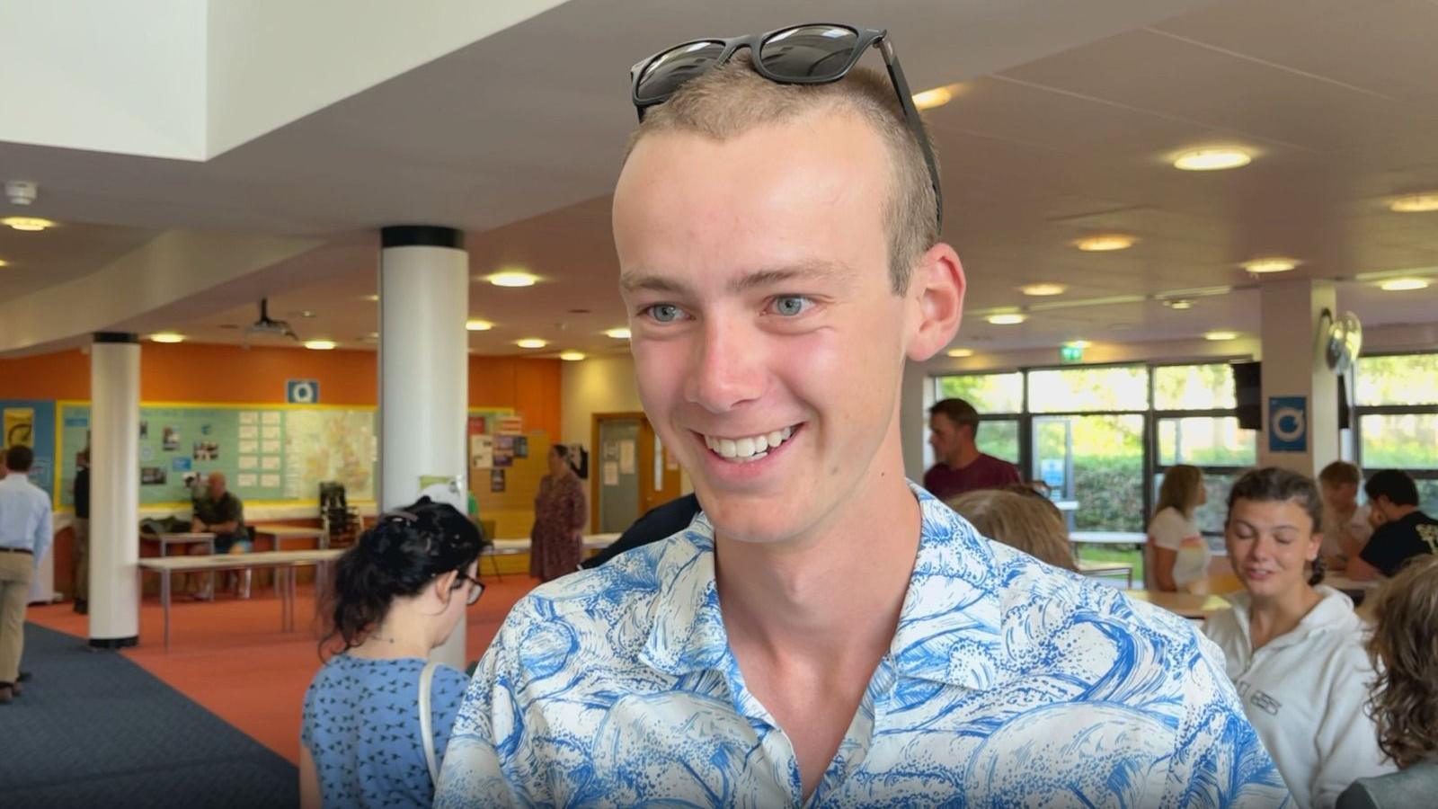 Sam Jones - a boy with short light brown hair smiling with a white and blue design shirt on, black sunglasses on his head, background of a school hall with people speaking behind