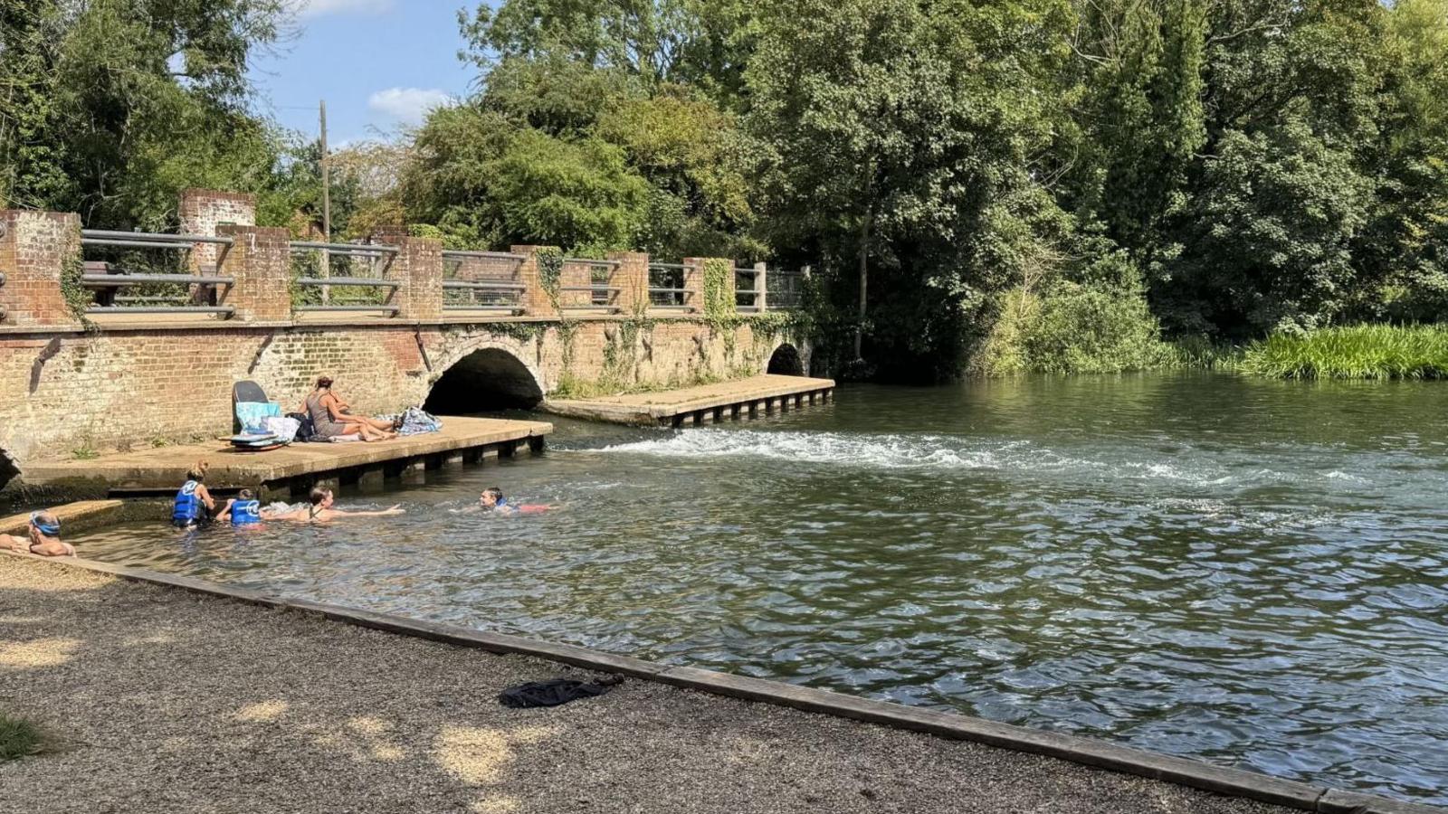 A brick bridge, with two arches, spans the river, with people on one platform next to an arch and swimmers in the water.
