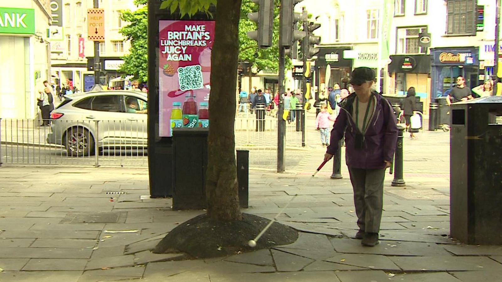 Terri Ballon, wearing a purple overcoat and grey cap, walks with her cane outstretched near an uneven surface where tree roots are pushing paving stones up to create a slant.