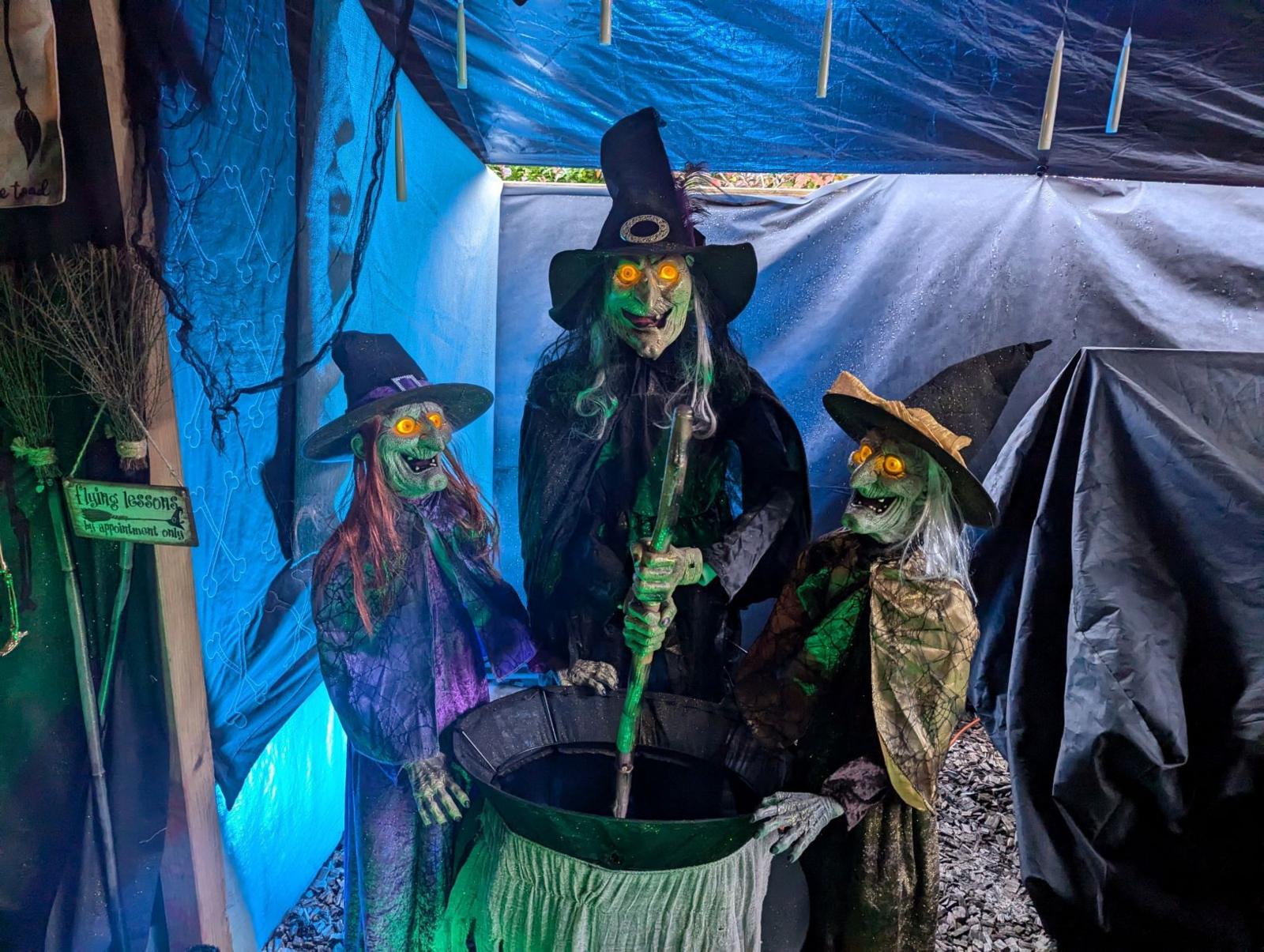 Three statues of witches around a cauldron. The witches have tall black hats and glowing yellow eyes. They are standing against a backdrop made of shiny black sheets, with blue and green lighting.