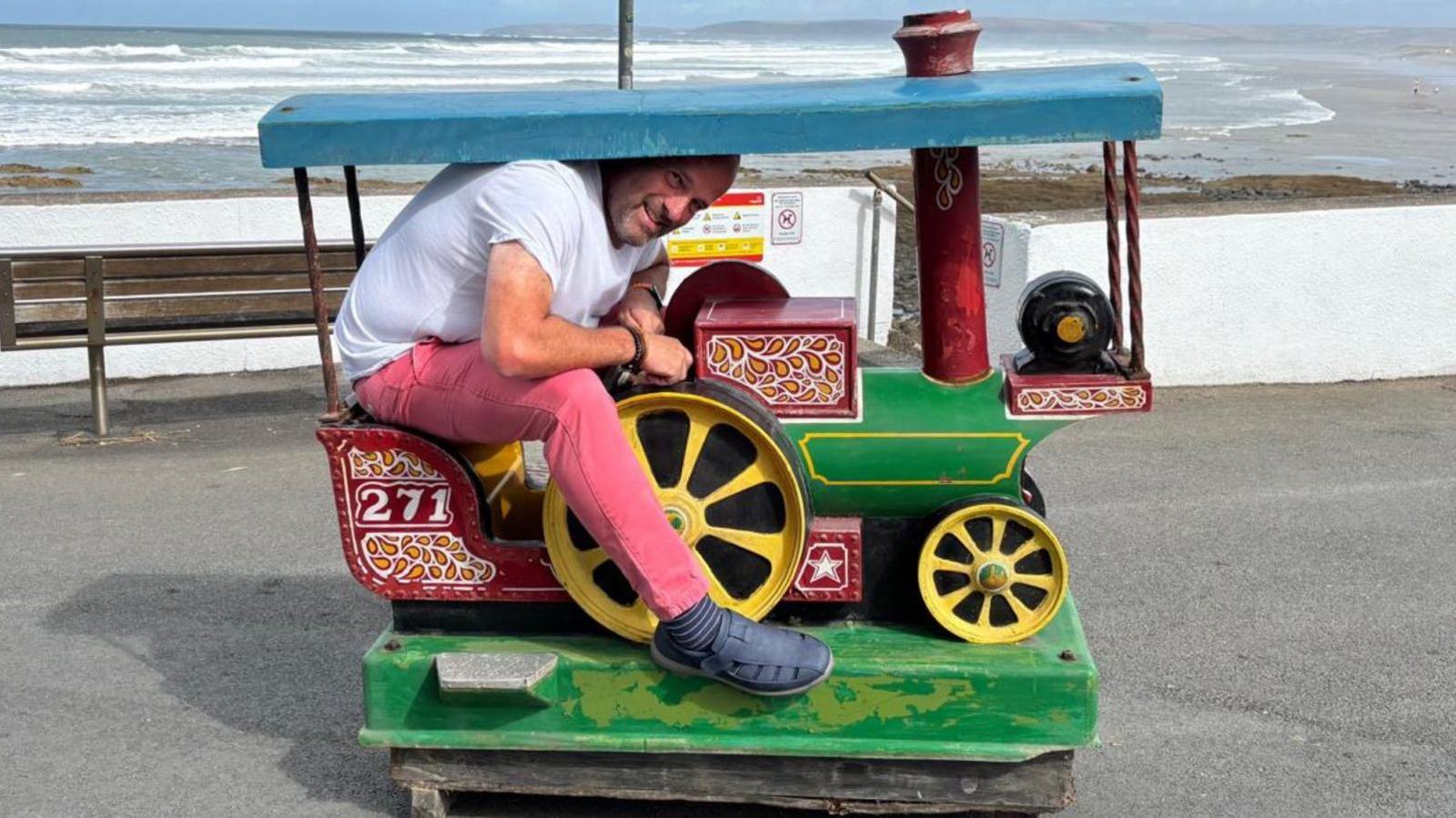 Adult Steve Dance is sitting on the seat of a toy steam engine. He has short brown hair and a beard and is wearing pink trousers and a white T-shirt with blue sandals. He is having to bend his head to fit under the canopy and looks too big for the ride. He is smiling and holding the steering wheel of the ride. It sits on the pavement and there is also an empty bench. The beach and sea are visible behind the pavement.