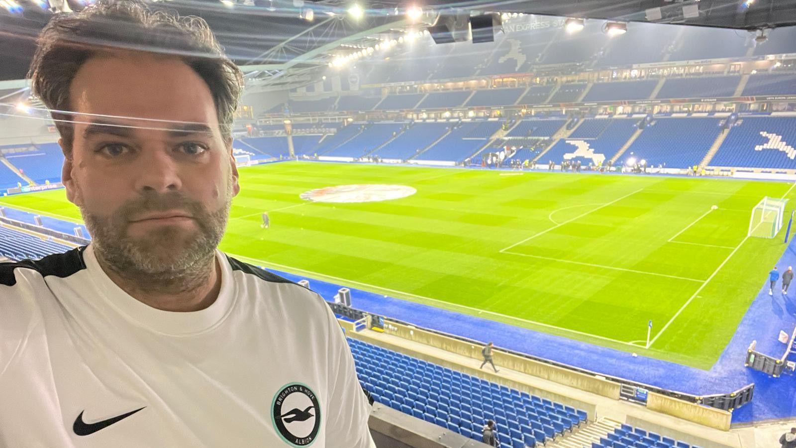 A selfie of Barry Hill at the Amex Stadium in Brighton. He is wearing a white Brighton & Hove Albion football shirt. The green pitch and blue stands are in the background. 