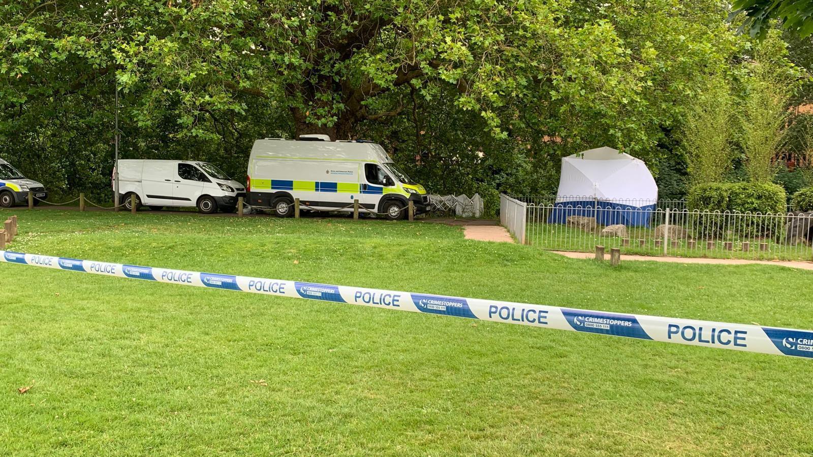 Police cordon in French Weir Park, Taunton. Police tape can be seen across the frame as well as police vans and a white forensics tent.