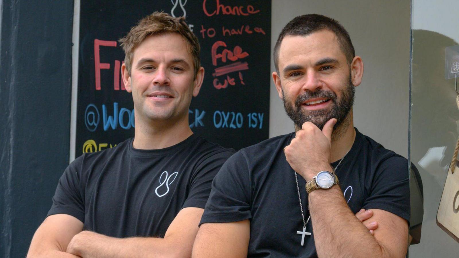 The Talbot brothers standing outside one of their salons. They wear black T-shirts, possibly the salon uniform. George has short brown hair. James' hair is more black, and he has a dark beard. He wears a cross around his neck.