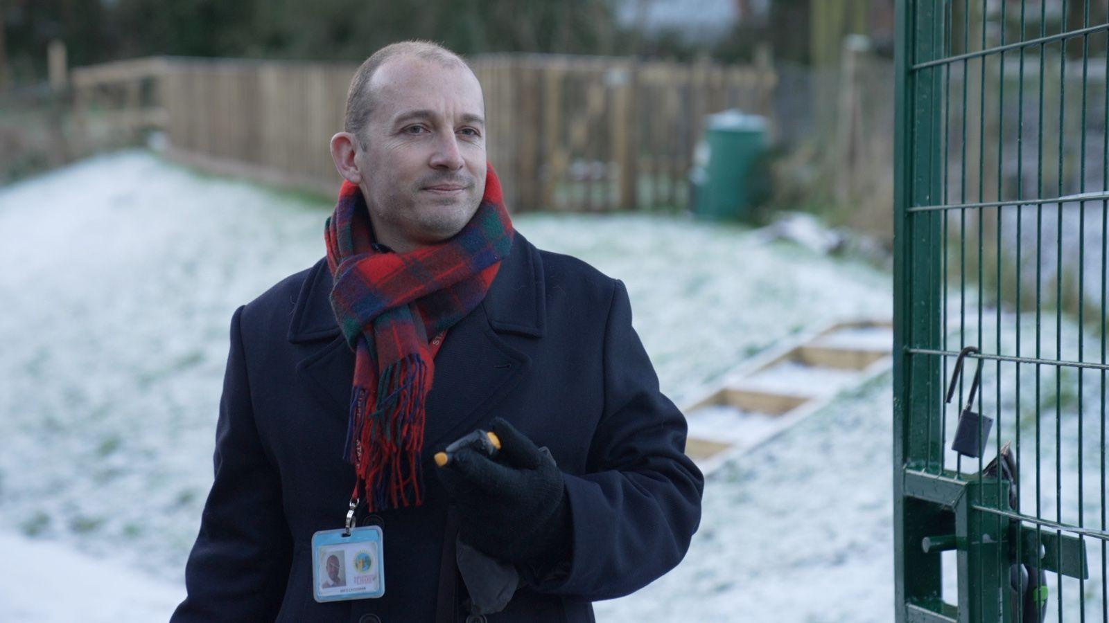 Head teacher Dan Crossman standing at the school gates. He is wearing a navy coat, checked scarf and there is snow on the grass behind him.