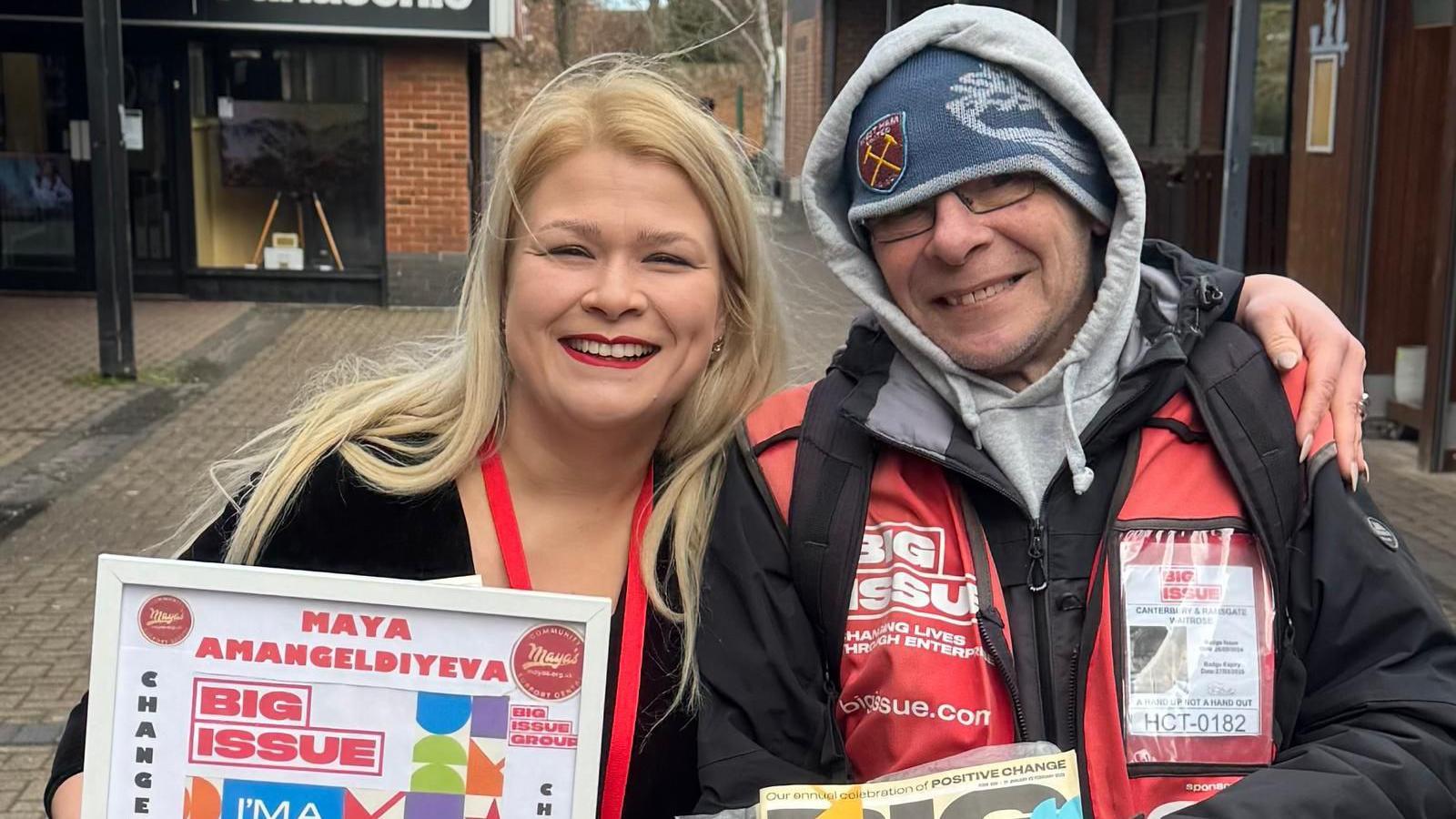 Maya, with long blonde hair, smiles and holds a certificate. She has her arm around Big Issue seller John, who is wearing a woolly hat and a hood.