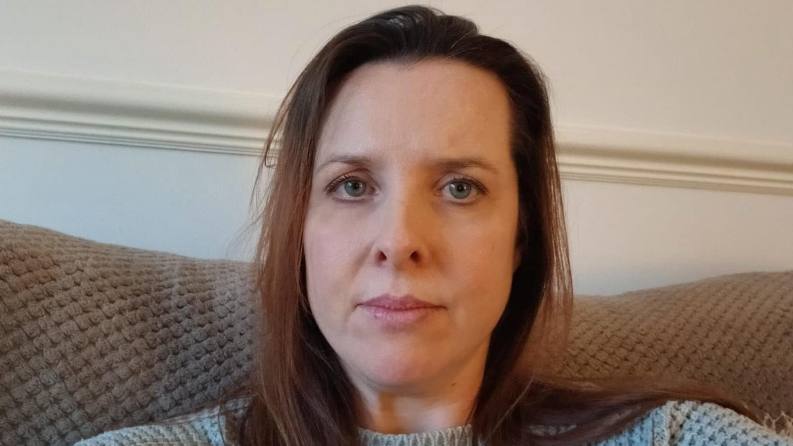 Jennifer, with long brown hair, sitting on a sofa staring directly into the camera