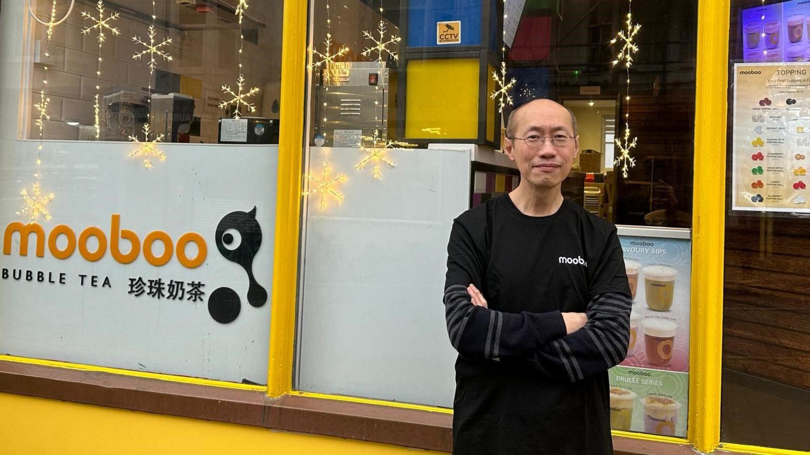 A bald man with glasses in a long-sleeve top standing outside a yellow bubble tea shop.