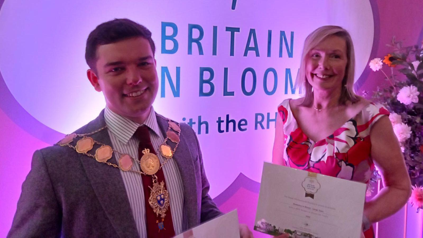 A man with brown hair is wearing a grey suit jacket, blue and white striped shirt, and red tie. He has a mayoral necklace on and is holding a paper award in his hands. Next to him is a woman with blonde hair in a white, red and pink floral dress. she is holding a paper award. they stand in front of a large banner reading "britain in bloom"