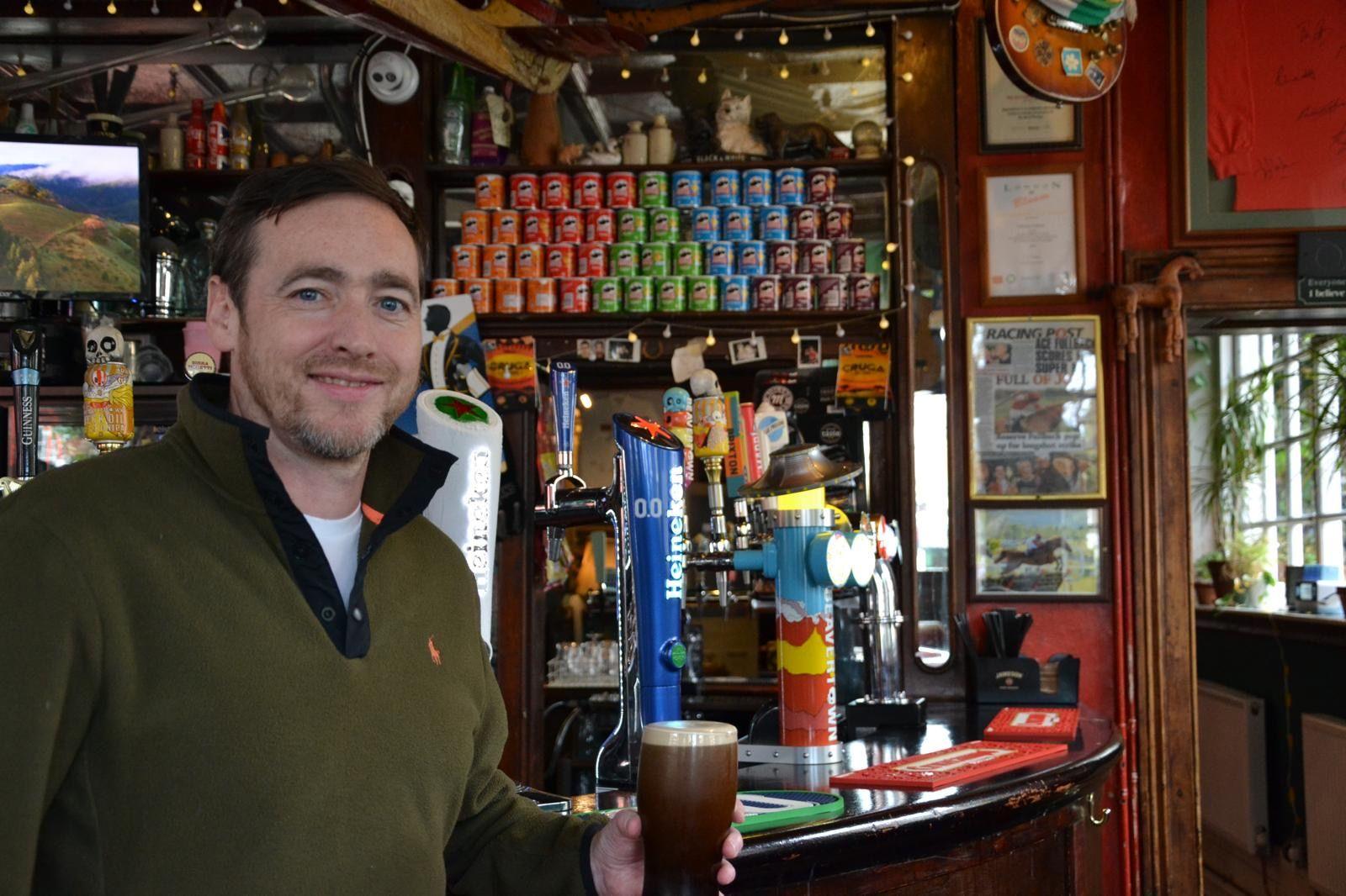 A man in a khaki fleece is holding a pint of beer and smiling at the camera. He is stood in front of a bar that has bar taps, tubs of Pringles and posters on the wall