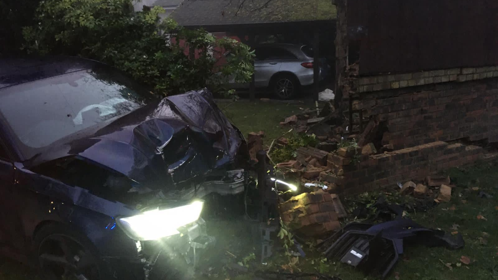 A blue car with a crumpled bonnet and one light on, partly in a hedge, next to a wall which has been broken. There is a hole the size of a car in the wall. Clumps of bricks surround the car. The airbag in the car is pressed up against the windshield. 