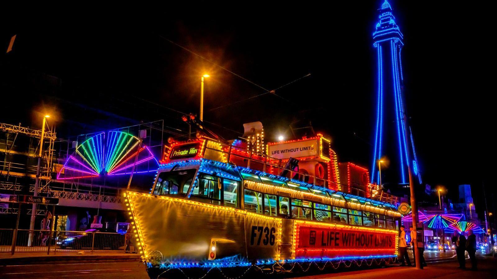 Illuminated tram 736 - HMS Blackpool - which looks like a ship on Blackpool's Promenade in front of Blackpool Tower in the evening which is lit up blue. The other illuminations can be seen in the background.