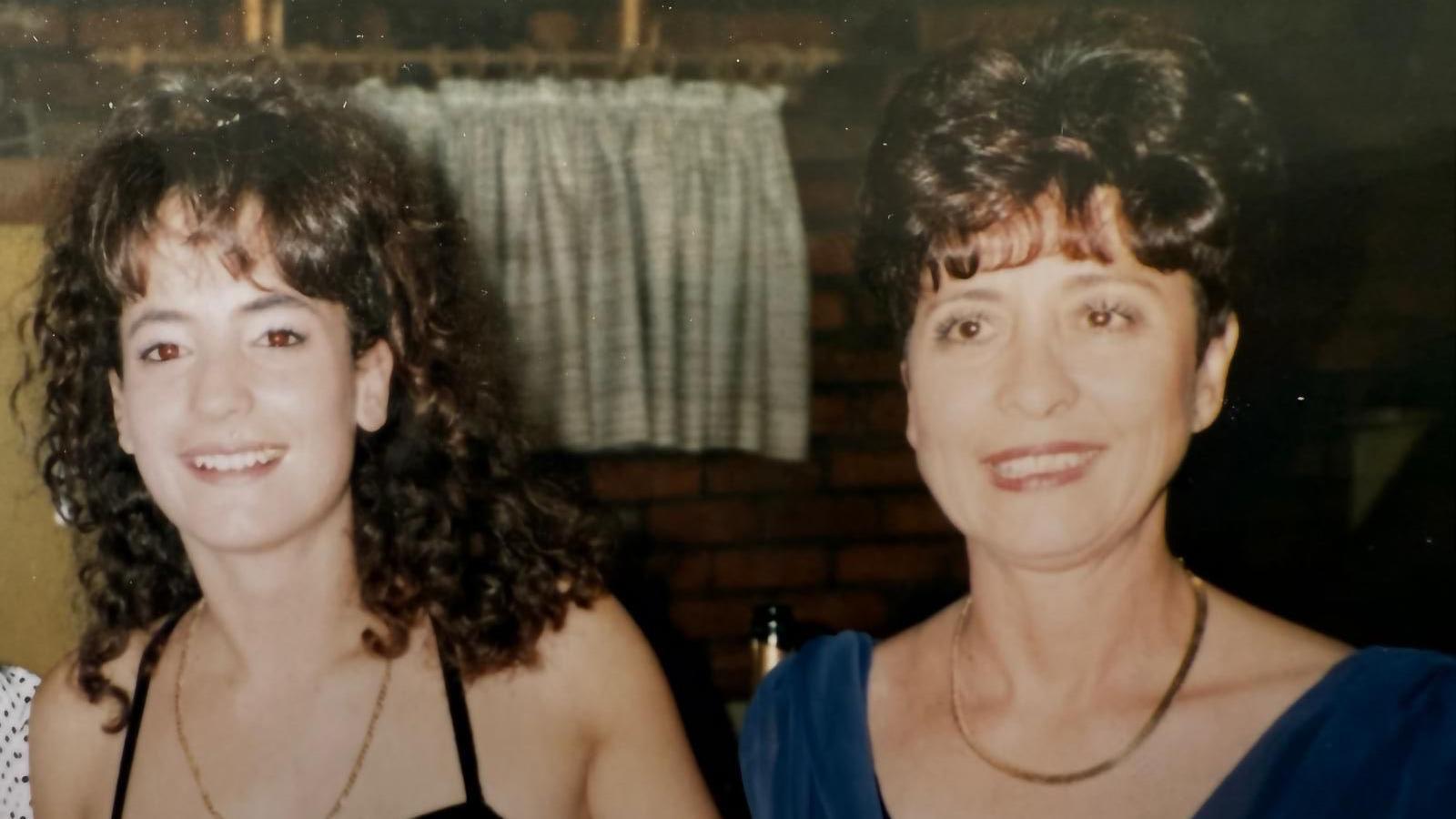 This is an old photo of Donna and her mum Carol. Donna is on the left with dark curly hair which comes down to her shoulders. She is wearing a black dress. Carol has a blue dress on and has shorter dark brown hair which is tied up into a bun.