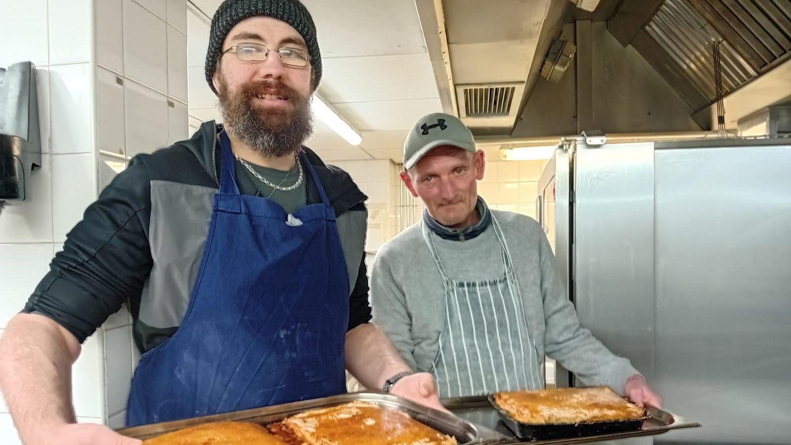 Two men in aprons hold up trays of pies