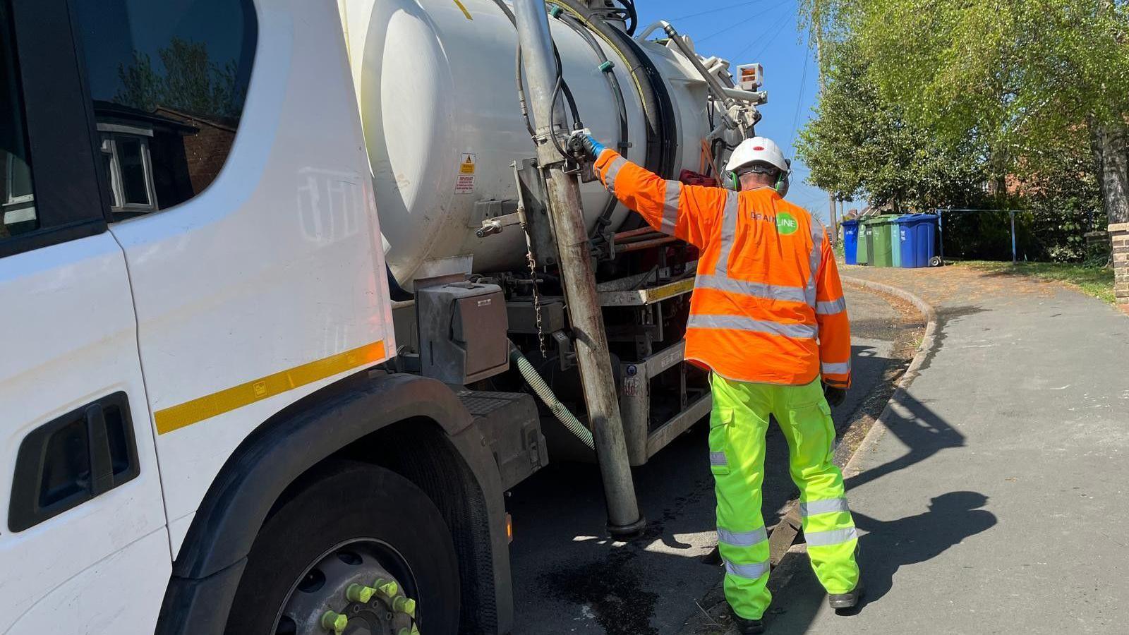 Worker wearing a high vis outfit and a helmet while cleaning a gully on a street, operating a drainage tanker. It is a sunny day.