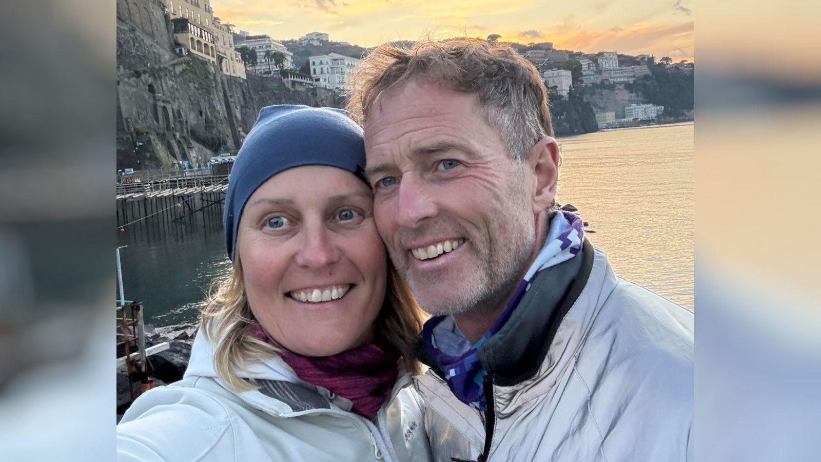 A man and woman are on holiday. They pose for a photo with a sea and sunset behind them. 