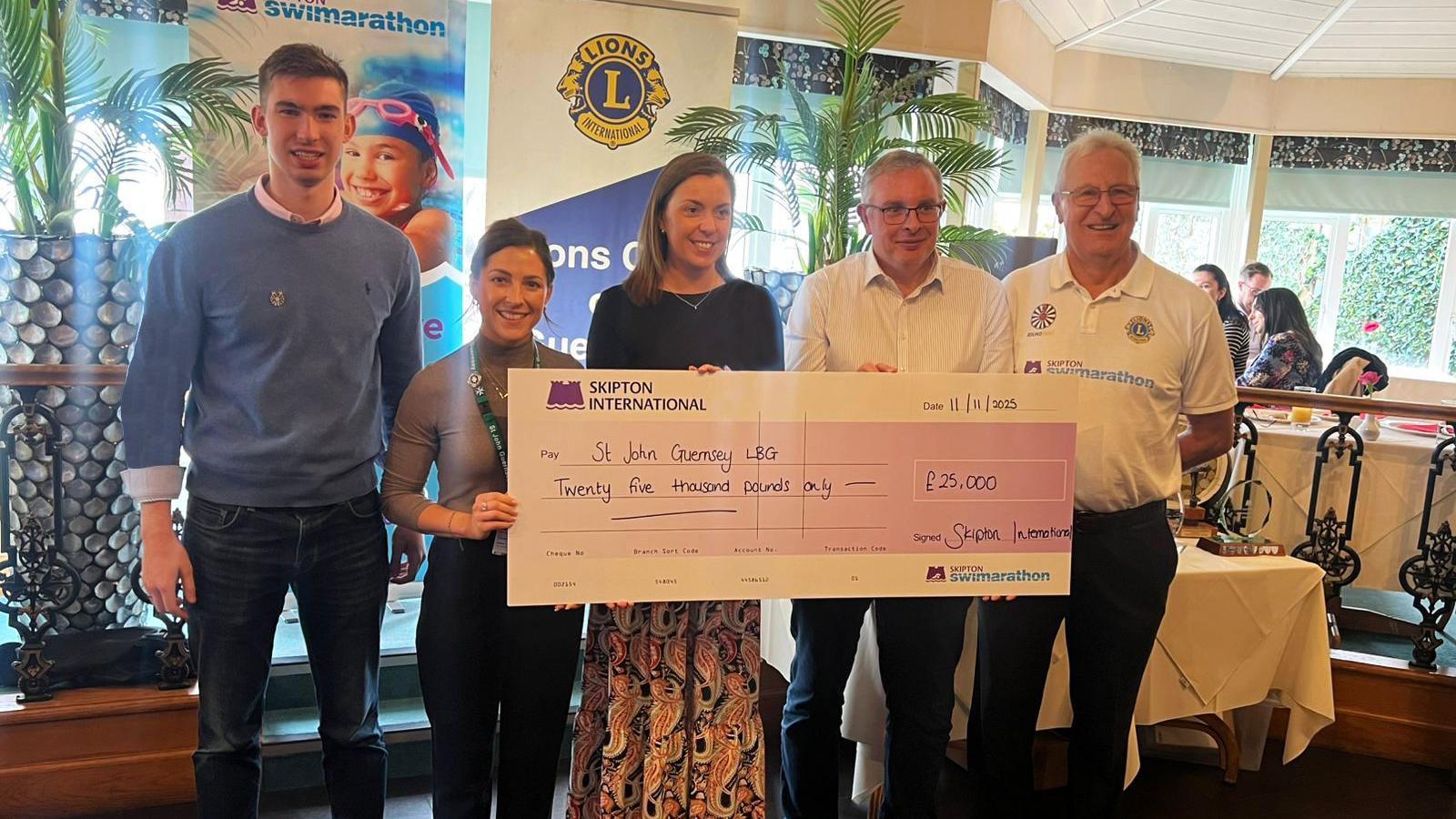 Five people stand in a line holding a large cheque made out to St John Guernsey LBG for the sum of £25,000. From L-R there is a tall man wearing a blue jumper, a shorter lady in a brown long sleeved top, a taller lady in black, and two older men in white shirts.