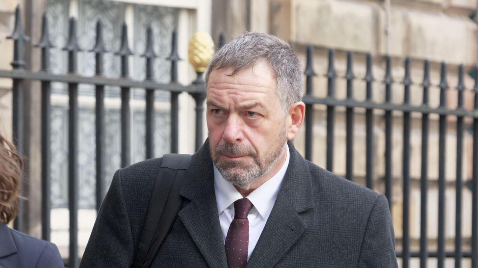 Tony Chambers, who has short grey hair and a grey beard, walks towards Liverpool Town Hall wearing a dark grey suit and sets his face in a serious expression