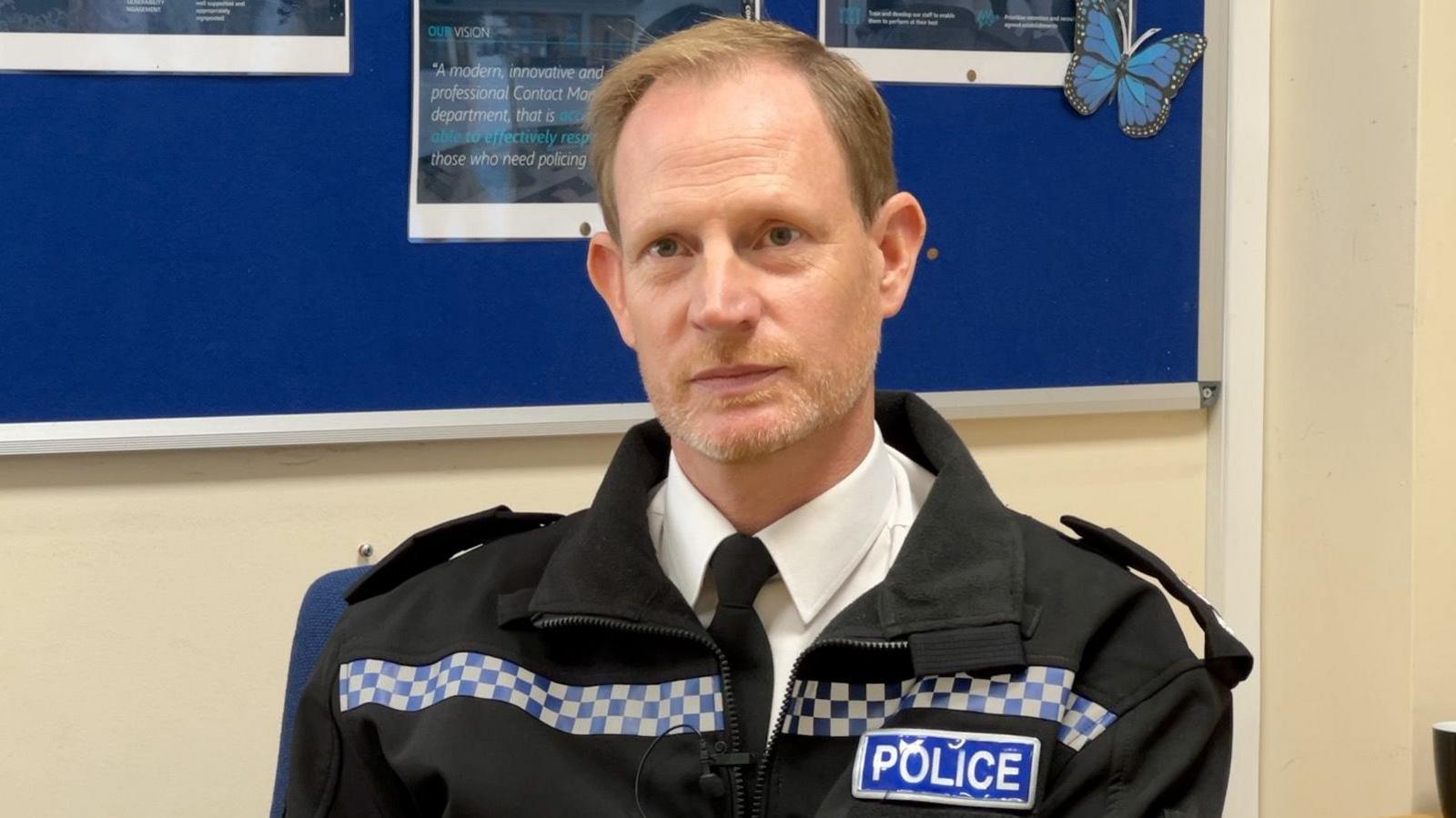 Chief Superintendent Simon Dodds has short light brown hair.  He is wearing police uniforn and is sat at a desk.
