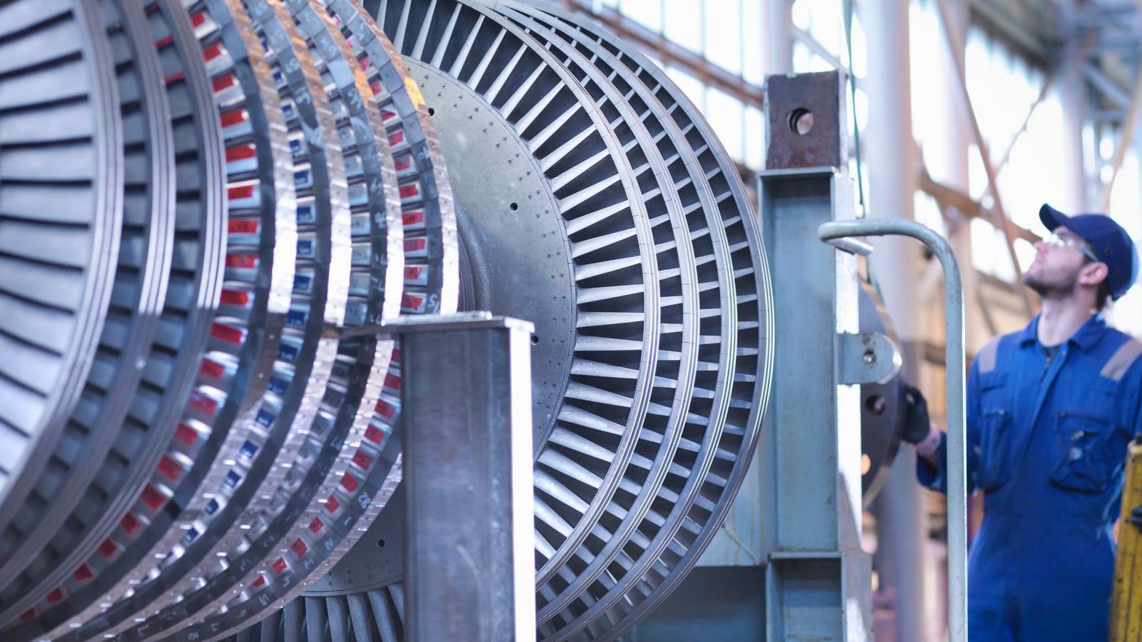 A man in a blue workers uniform is standing to the right of a steam turbine in some kind of electricity plant. He is looking up at the turbine, monitoring it, while wearing safety goggles and a blue cap. 