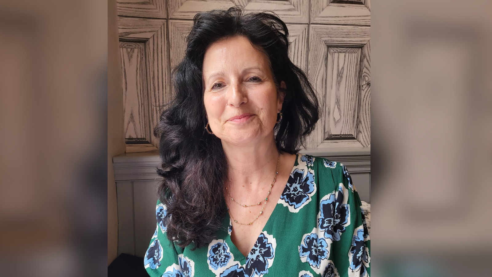 A woman with long, wavy dark hair. She wears a green top with blue flowers, and a gold necklace and earrings.