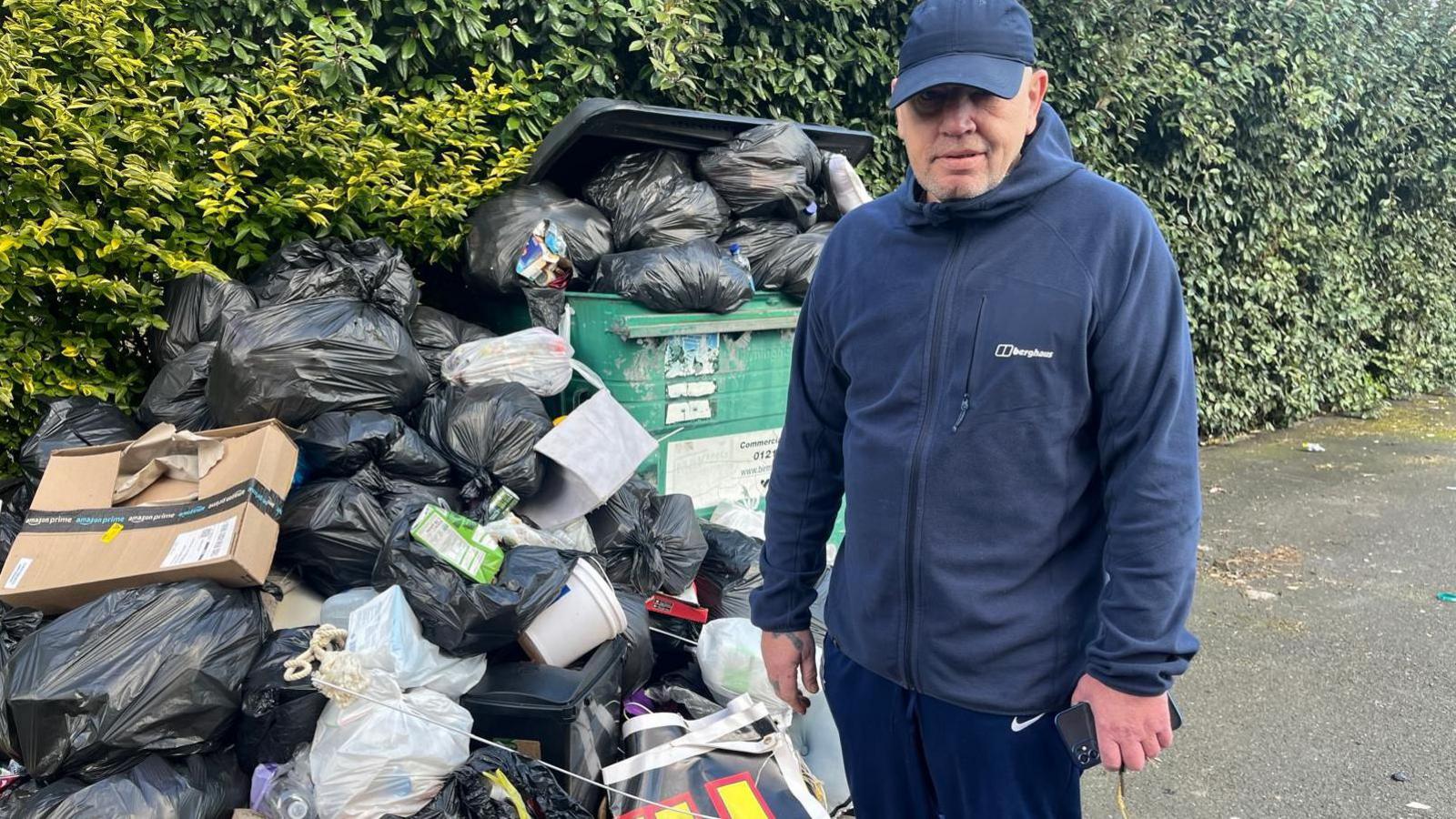 Teddy Smith standing next to black bin bags on the street next to an overflowing industrial bin. He's wearing a blue fleece, blue tracksuit bottoms and a blue cap.