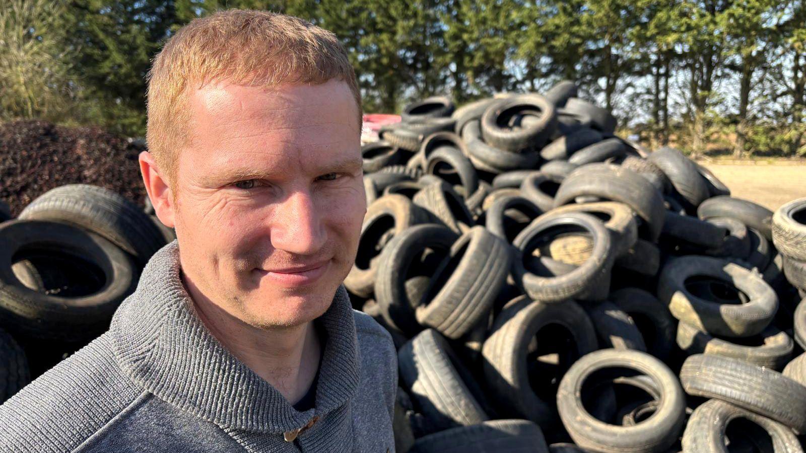 It is a sunny day and Elliott Mason, who has short fair hair smiles in the foreground, wearing a grey zipped jumper. Behind him is a large pile of waste tyres and trees line the background.  