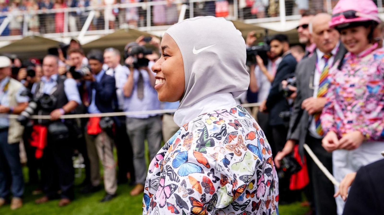 A woman wearing a hijab and a colourful horse-riding top. There is a crowd in the background. 